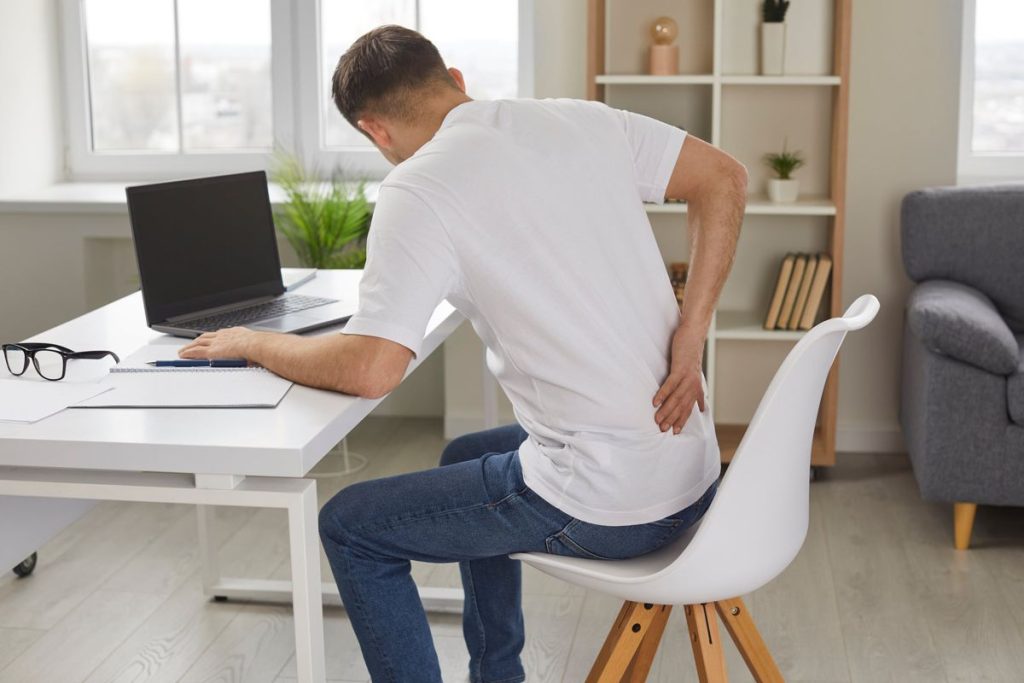 Young man experiencing lower back pain while working at desk, indicating poor posture or prolonged sitting.