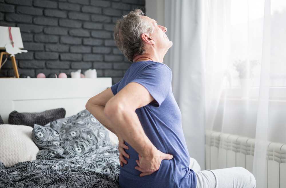Older man holding lower back in pain while sitting on bed, showing symptoms of chronic back discomfort.