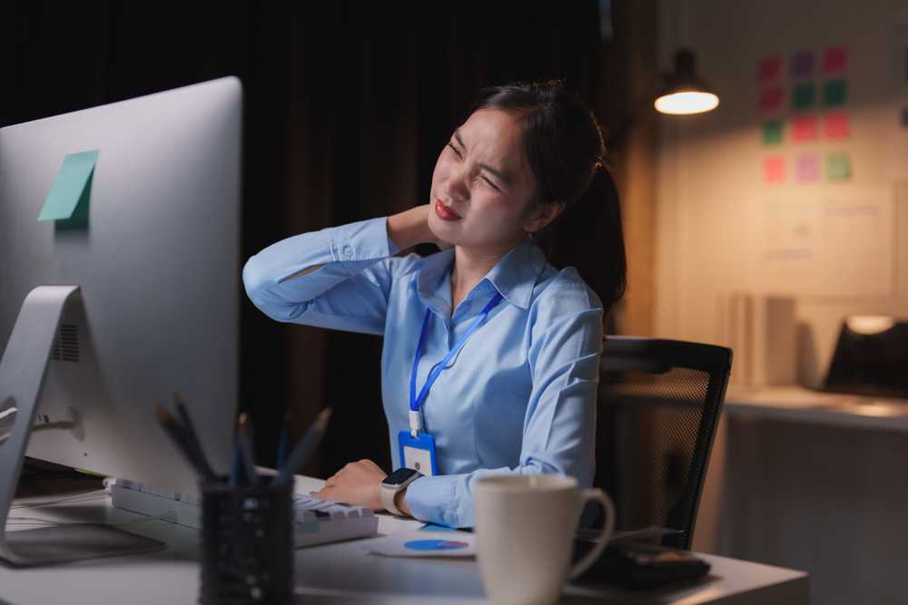 Office worker experiencing neck pain from poor posture while working at a computer late at night.